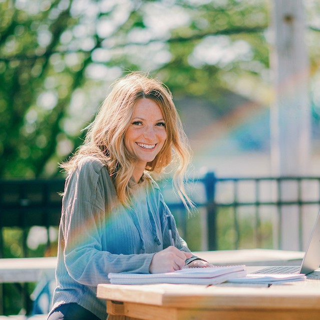 Online: Woman sitting at a table outside on a sunny day, smiling at the camera.