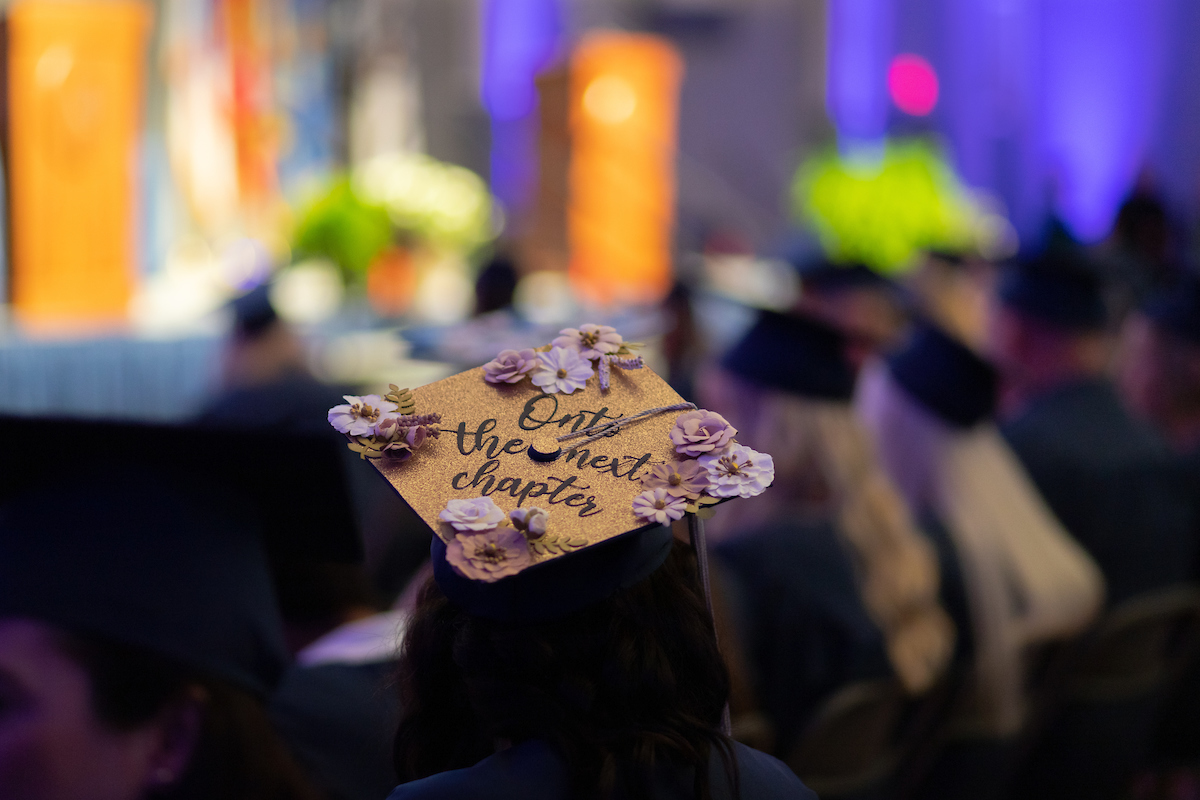 Back view of graduate during commencement, displaying "Onto the next chapter" on her graduation cap.