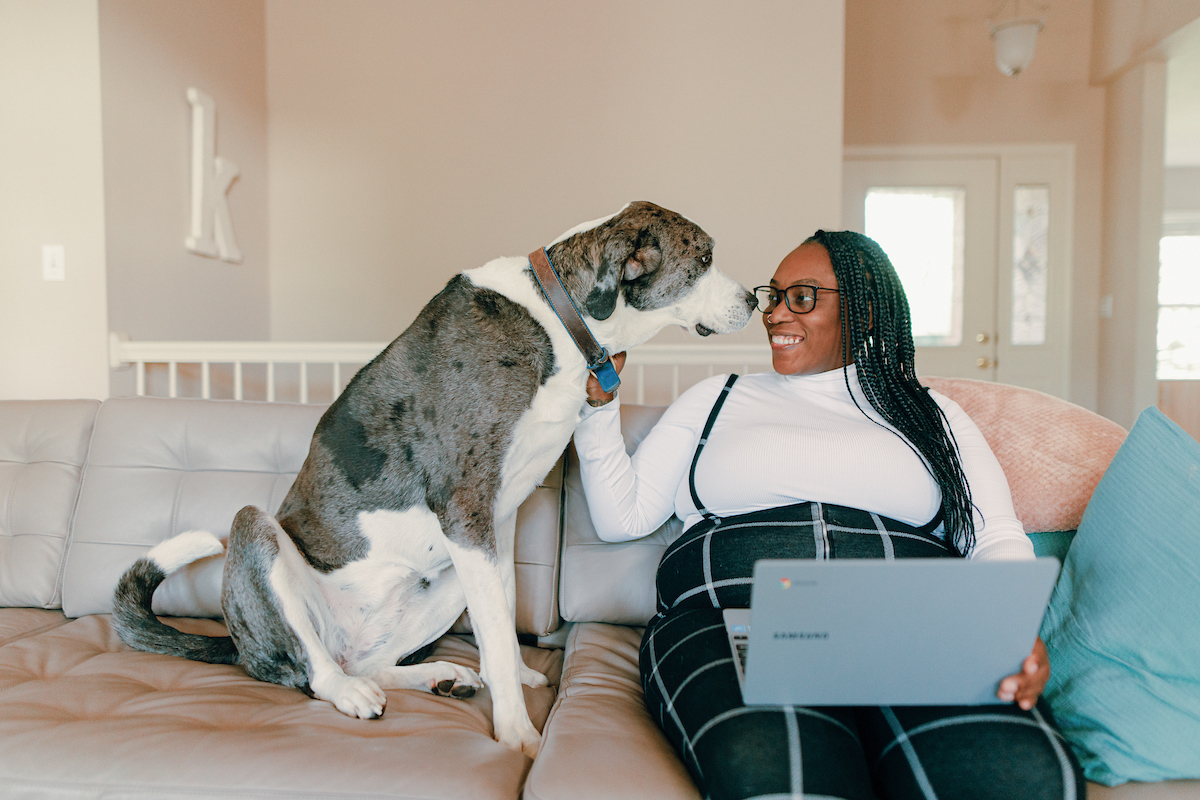  Adult student sitting on couch with a laptop in her lap and a large dog snuggling up next to her.