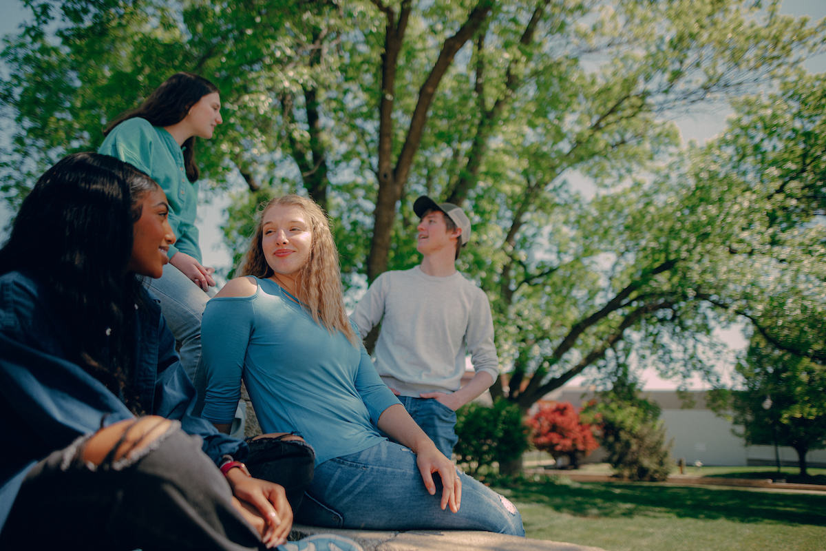 Four students engaged in conversation in front of a large tree on main campus in the spring.