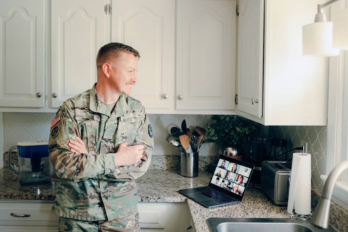 Man in military uniform standing at the kitchen counter reviewing content on his laptop.