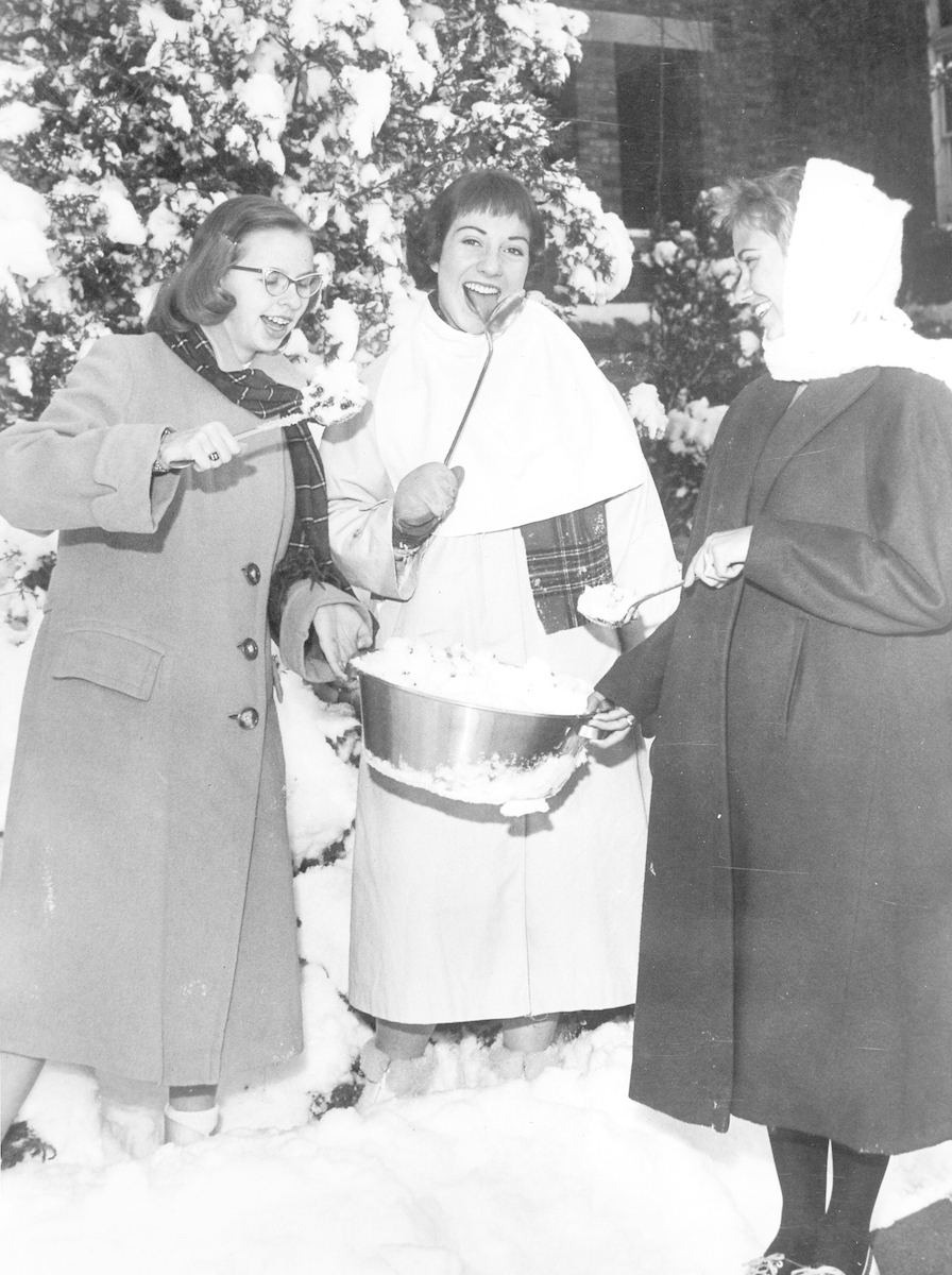 Historical image of three women wearing winter coats, eating "snow" cones from the snow.