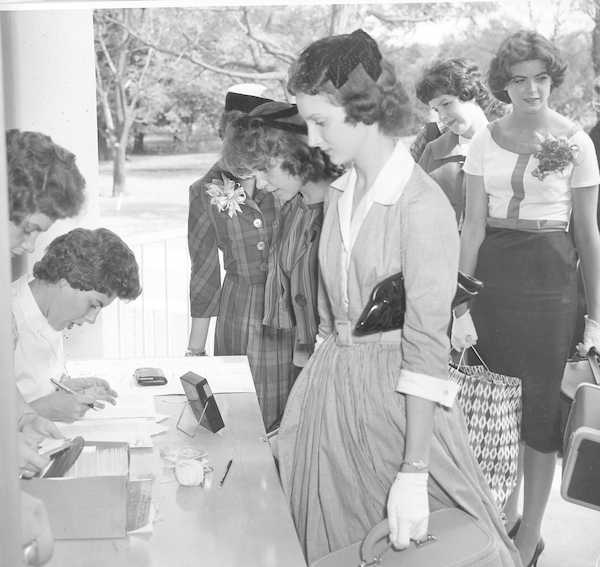 Historical image of women standing in a line, wearing dresses, hats and corsages, checking in at a table.