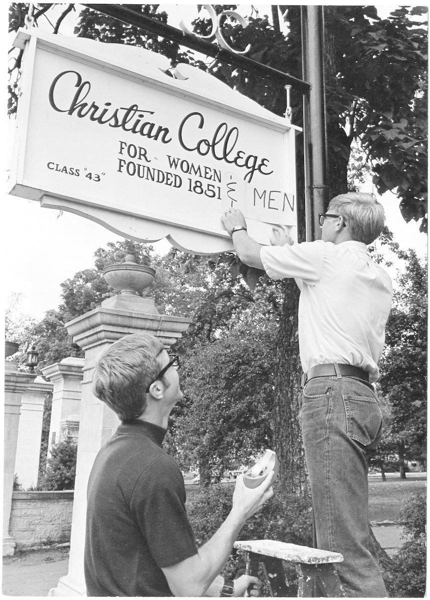 Historical image of two male students adding a sign that says "& Men" to the "Christian College For Women" sign on the main campus.