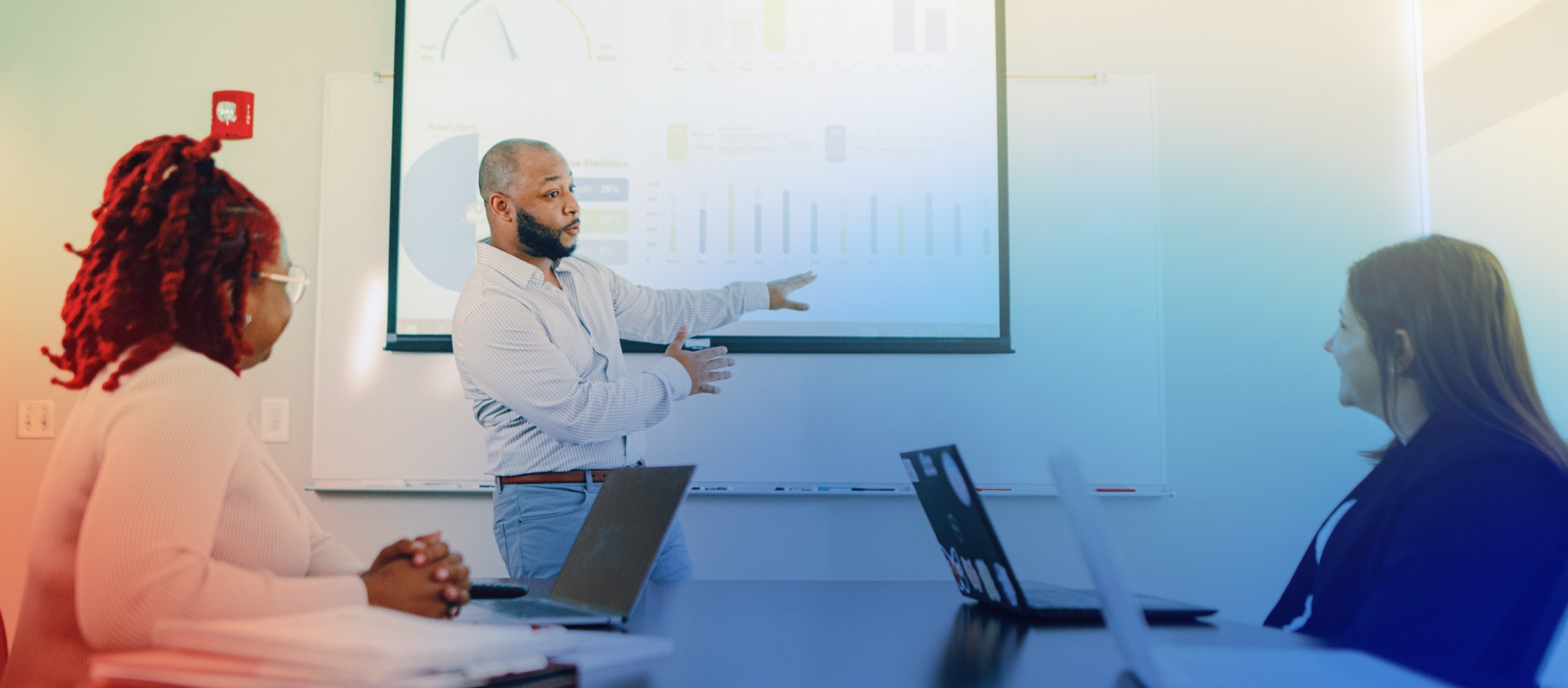 Man standing in front of white board, explaining contents to two women, seated.