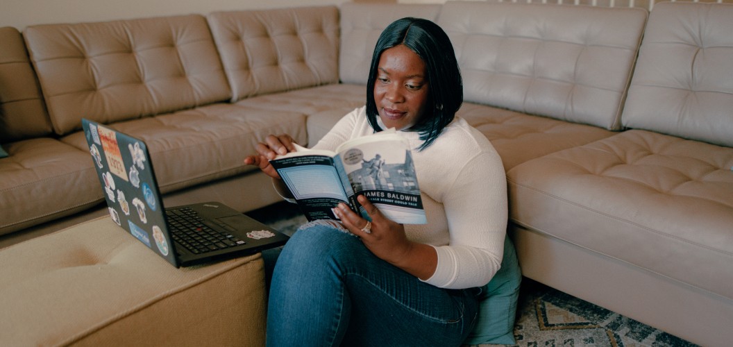 Woman sitting on floor, leaning against a pillow and couch, with a laptop in front of her while she leafs through a book.