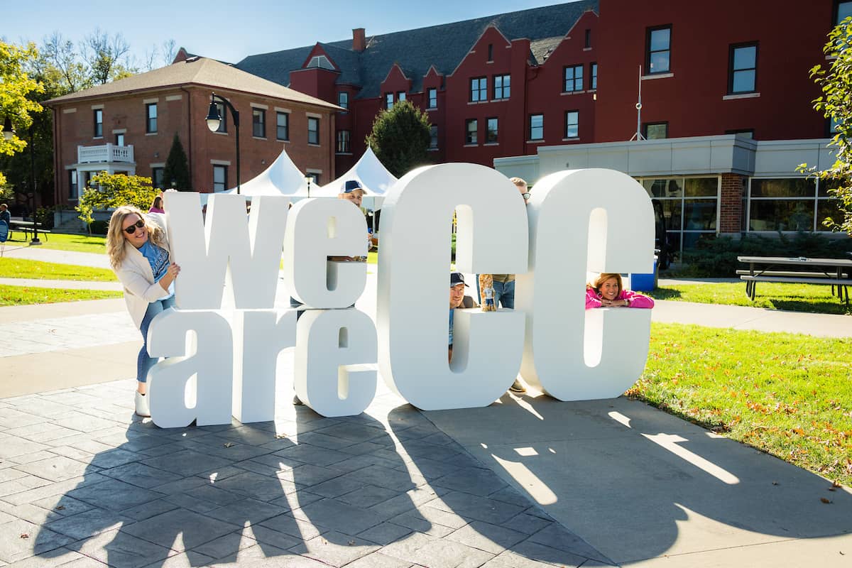 Students peaking through the We are CC sign, with the sun causing a shadow of the sign on the sidewalk.