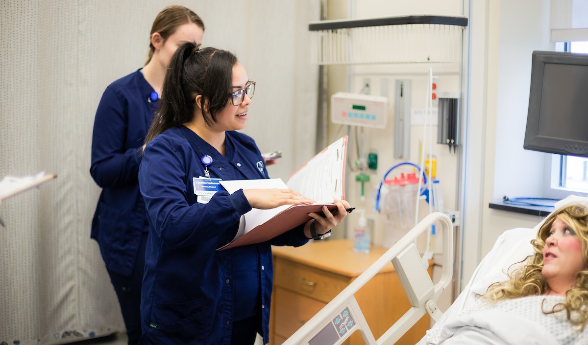 Nursing students stand by a "patient's" bedside talking, while reviewing charts.