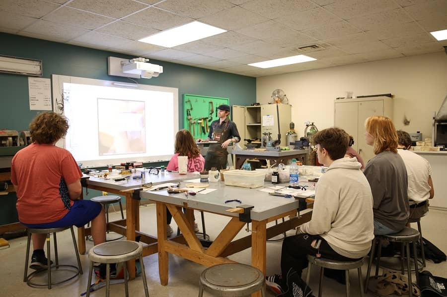 Six students sit on stools around work tables, looking at information on the board, with the camp instructor standing.