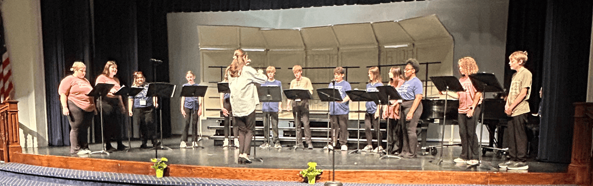 Choir Fest campers stand on stage with their music stands in front of them, performing a song while the instructor leads them.