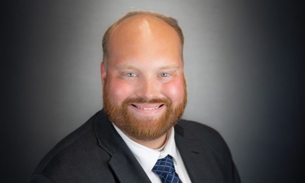 Headshot of Nick Stegmann, wearing a dard wuit, shite shirt and blue tie with a criss-cross pattern, smiling at the camera.