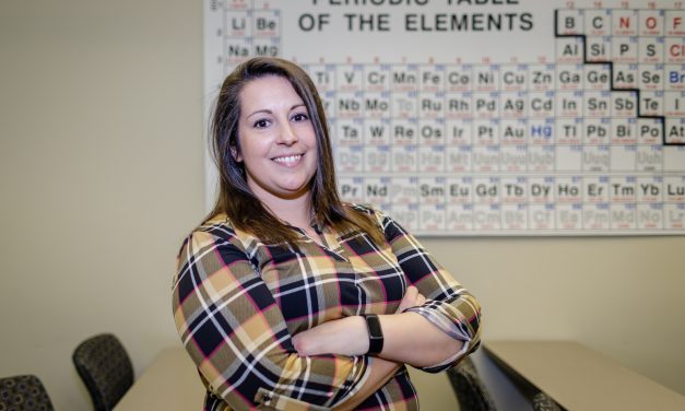 Dr. Brittany Hagenhoff, wearing a gold, black, white and pink plaid shirt, stands in front of a wall size Periodic Table.