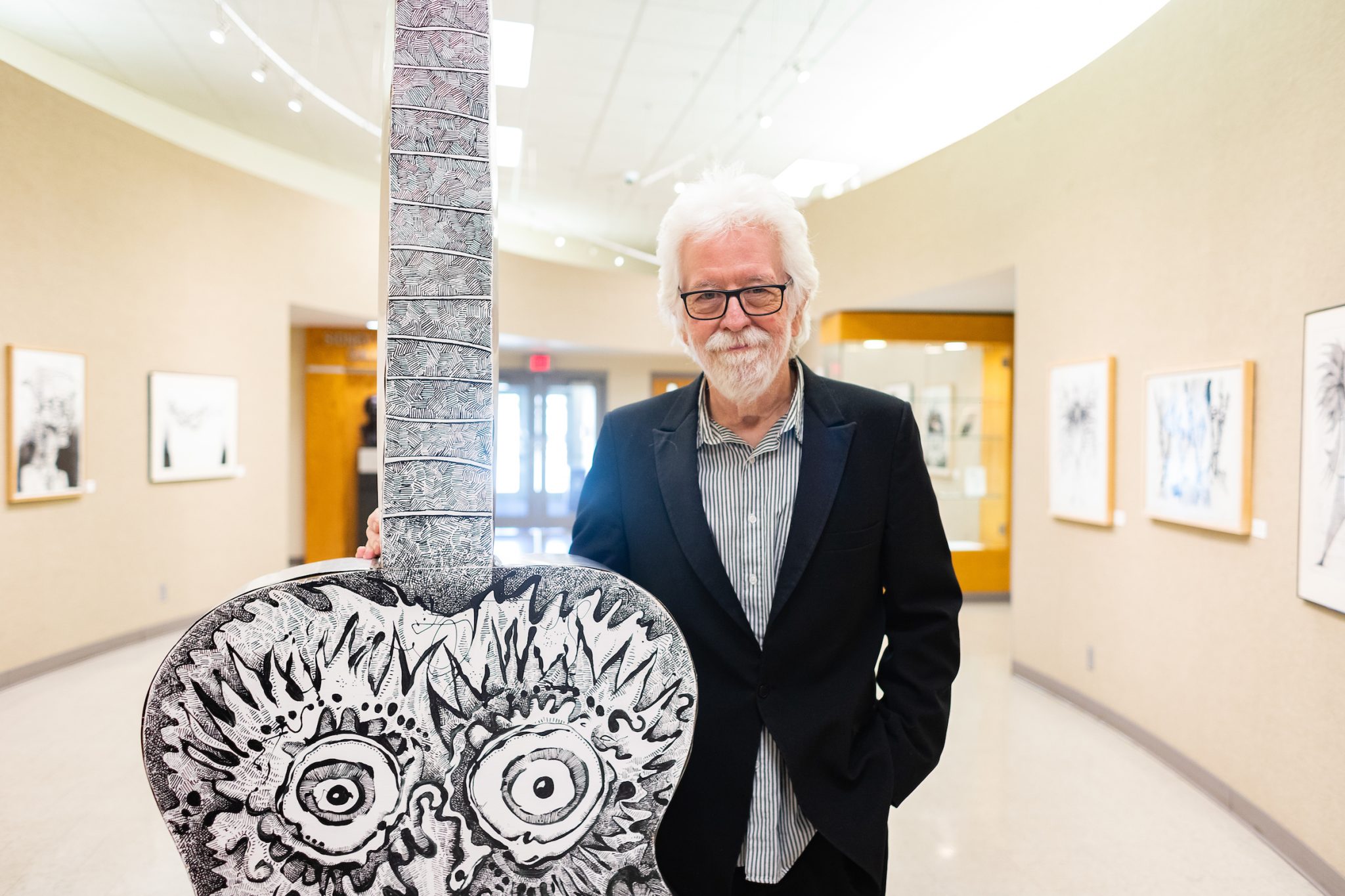 Art Professor Mike Sleadd poses next to a piece of his artwork, depicting an owl on a giant guitar.