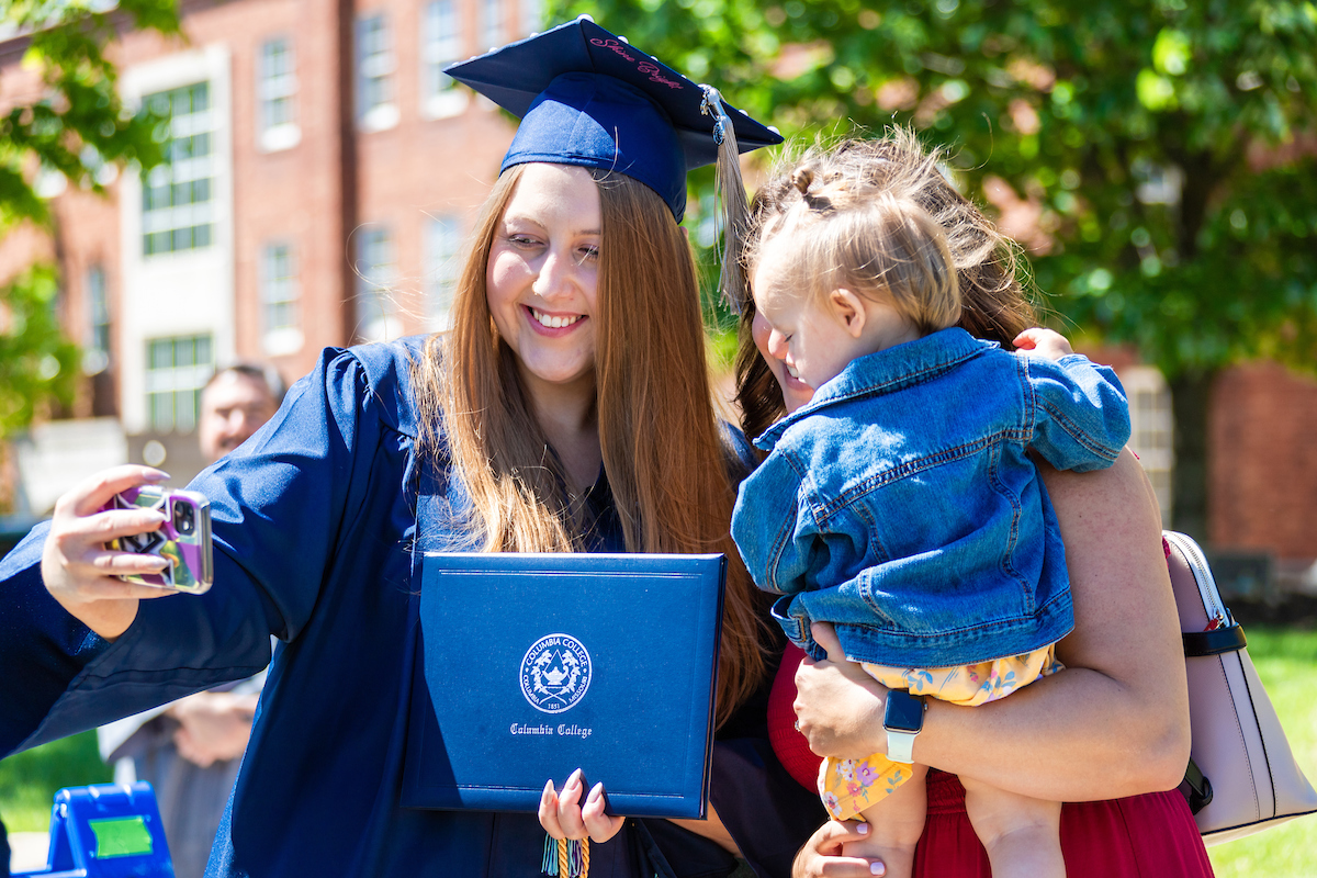 A graduate, in full regalia, standing next to a woman holding a young child, taking a selfie.