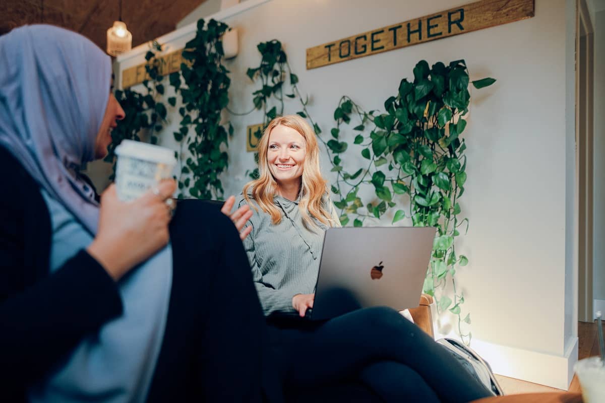 Two female students having a conversation, with one student holding a laptop and another wearing a head scarf and holding a cup of coffee.