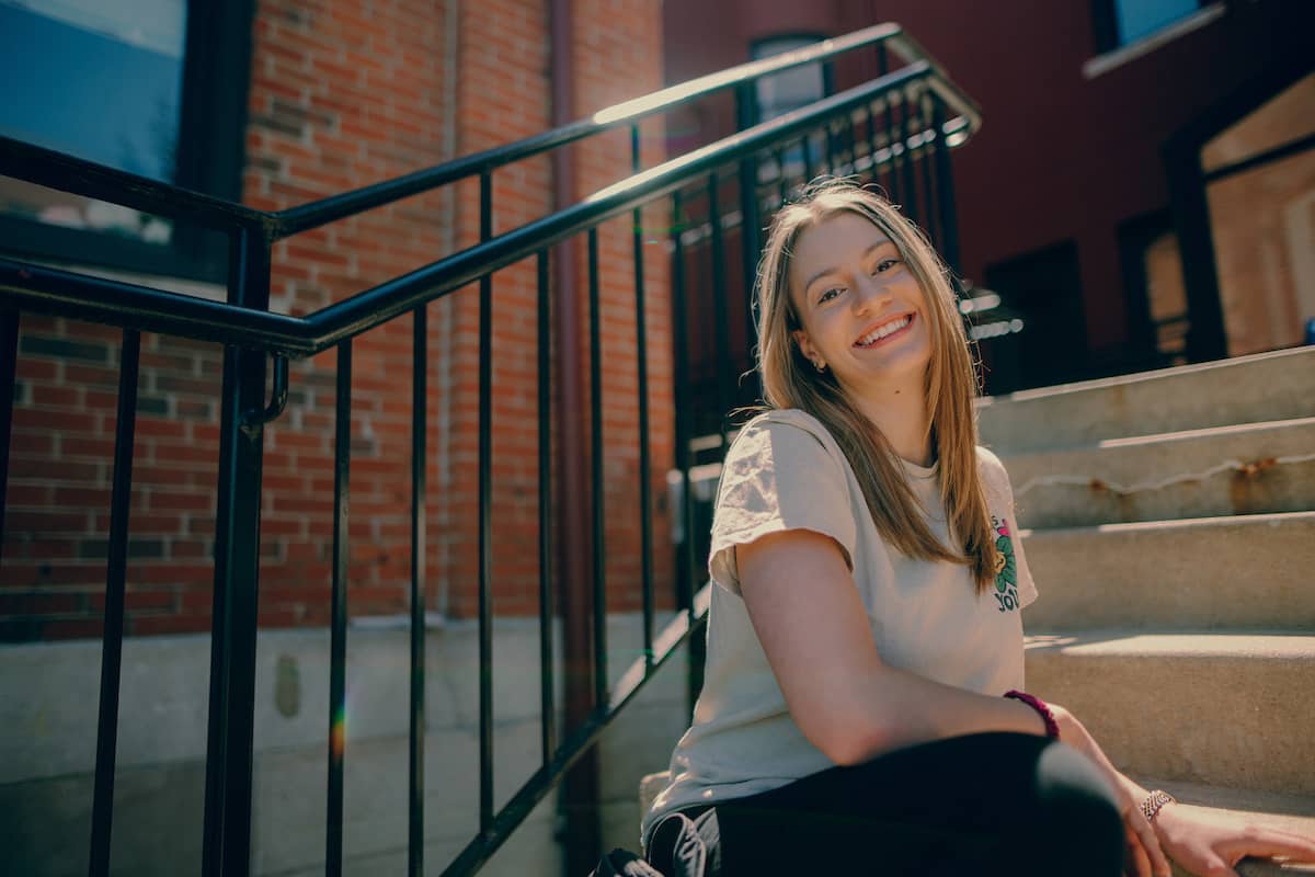 Female student sitting on steps on main campus, turned slightly to smile at the camera.