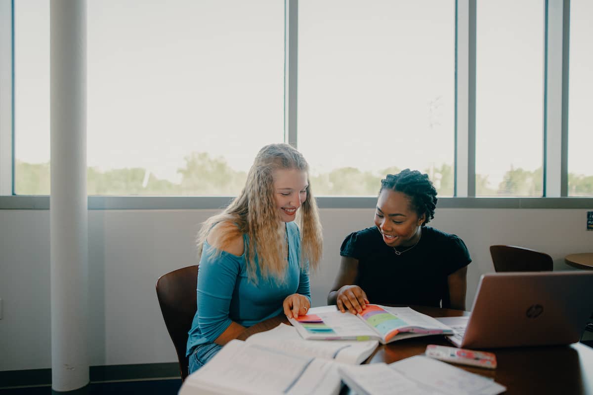 Two female students sitting at a table in front of a bank of windows, reviewing a book together as part of the Aspire Business Clinic consulting team.