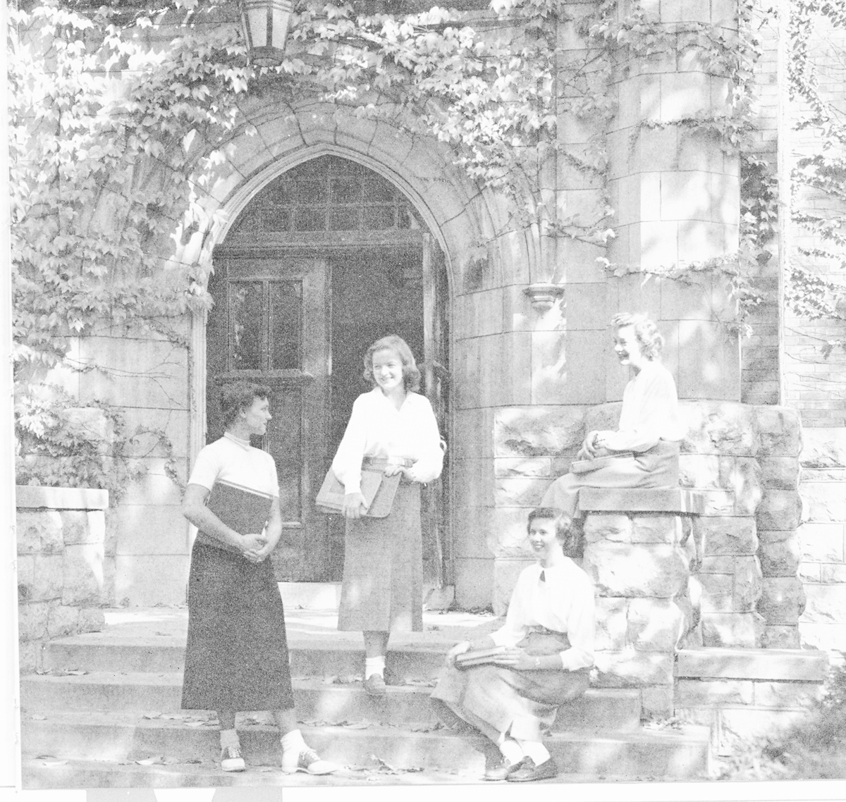 Historical image of two female students walking through the arched stone entryway of a main campus building.