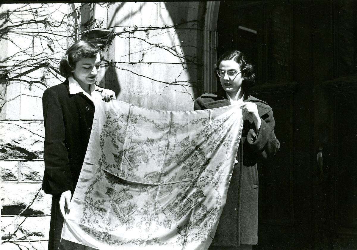 Historical image of two women, standing and holding a large scarf filled with design.